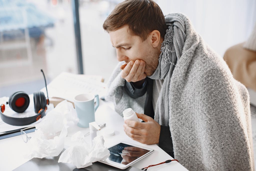 Man wrapped in blanket feeling unwell at home, surrounded by tissues and medication.