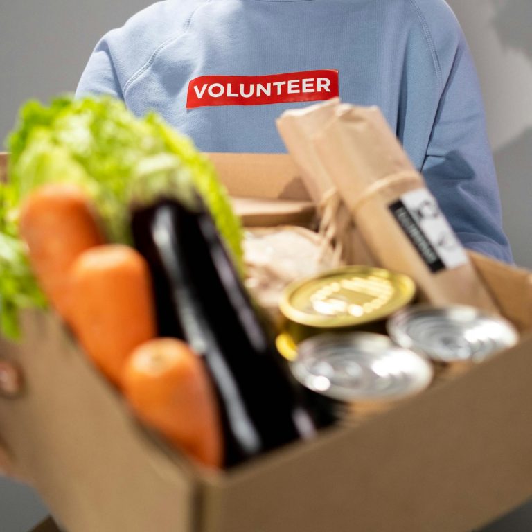 Smiling woman holding a box filled with various vegetables and canned goods during a volunteer drive.